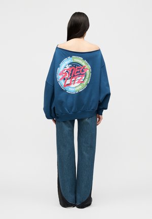 Kobieta o długich ciemnych włosach nosi niebieską, dużą bluzę z odkrytymi ramionami i szerokie niebieskie jeansy, stoi tyłem na białym tle.