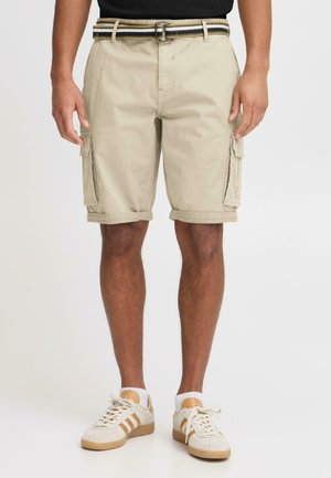 Uomo che indossa pantaloncini cargo beige con orli arrotolati, cintura a righe, calzini bianchi alla caviglia e sneakers casual beige, in piedi davanti a uno sfondo semplice.