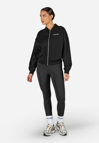 Melnā rāvējslēdzēja bomber jaka ar ribotām piedurknēm, kombinēta ar augsto jostasvietu melnām legingām. Iekļauj neitrālu toņu sporta apavus un baltas zeķes.