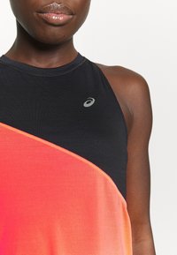 Nahaufnahme einer Person, die ein ärmelloses, schwarzes und orangefarbenes Sporttop mit einem kleinen silbernen Logo auf der Brust trägt.