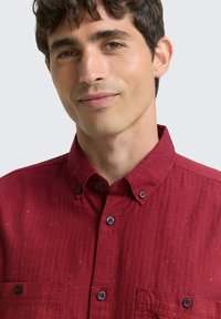 Chemise rouge à boutons avec un tissu texturé, dotée d'un col pointu, d'une poche poitrine et de boutons contrastants. Présence d'un petit motif tacheté.