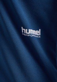 Blau strukturiertes Gewebe mit weißem "hummel PERFORMANCE"-Logo, das leichte Falten und Schatten zeigt.