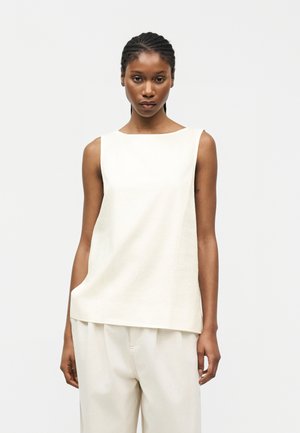 Vrouw met gevlochten haar, gekleed in een wit mouwloos topje en losse witte broek, tegen een eenvoudige lichte achtergrond.