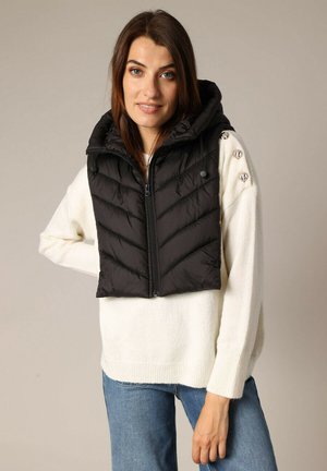 Vrouw in een witte trui met decoratieve knopen op de schouder, gedragen met een korte zwarte gewatteerde bodywarmer met capuchon, poserend met één hand op haar heup.