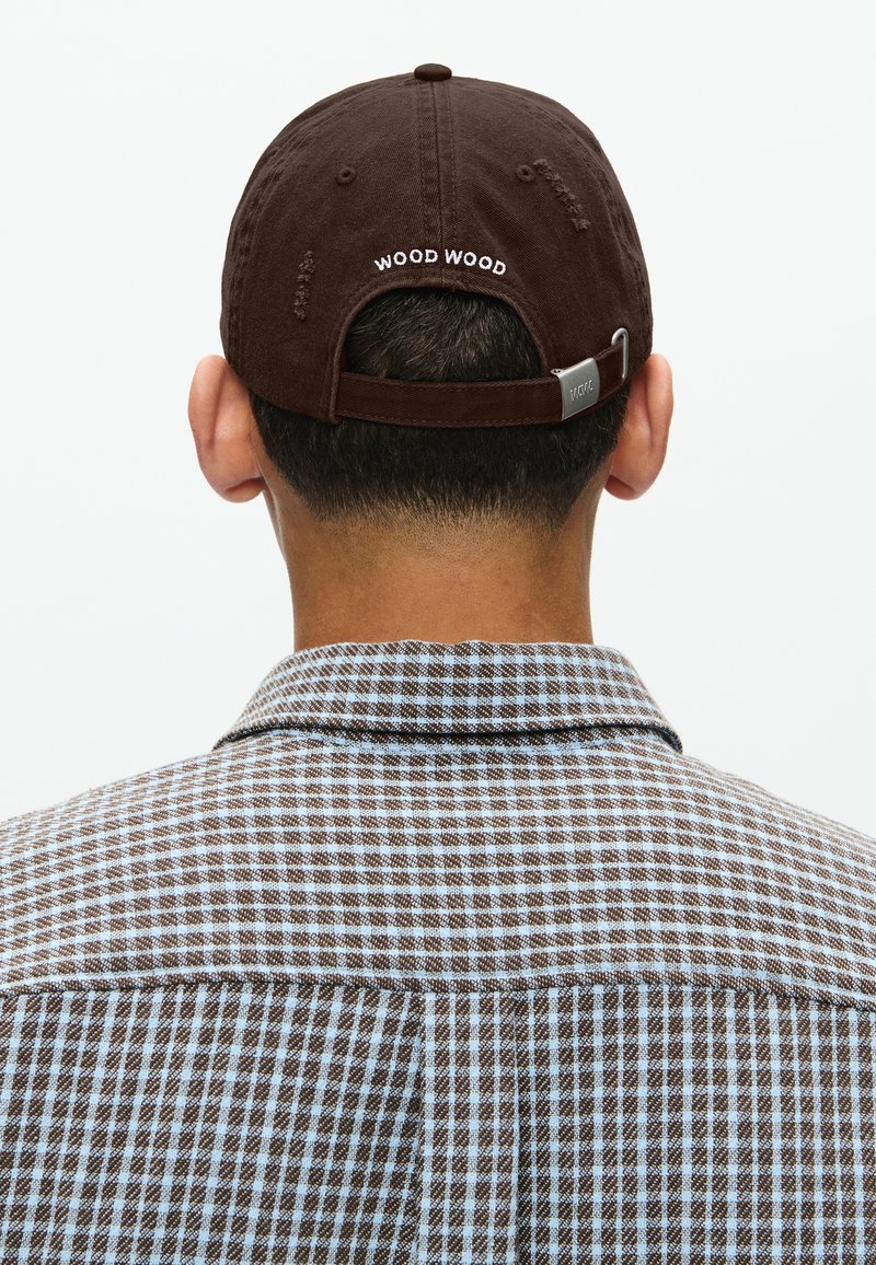 Cappellino da baseball marrone con la scritta "WOOD WOOD" ricamata sul retro, indossato sopra una camicia a quadri marrone e blu. Clip metallica sulla cinturino del cappellino.