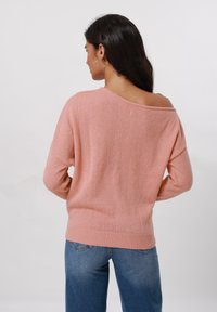 Un pull rose doux avec un large décolleté asymétrique et des poignets côtelés. Présente un tricot texturé, associé à un jean en denim bleu.
