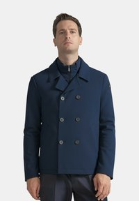 Uomo in piedi che indossa un giubbotto doppiopetto blu navy con una cerniera sotto, rivolto in avanti su uno sfondo semplice.