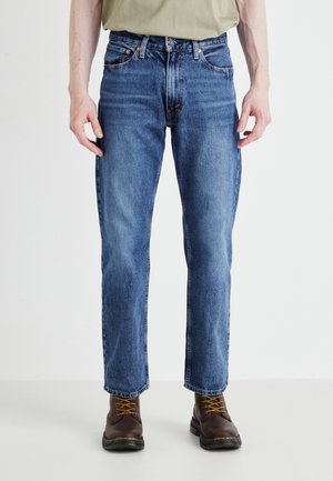 Personne portant un jean bleu droit avec des bottes marron à lacets, debout devant un fond clair et uni.