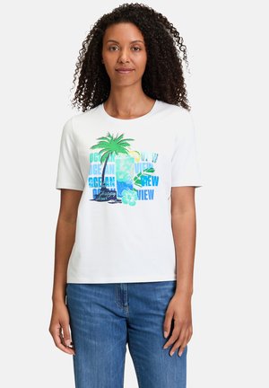 White short-sleeve t-shirt featuring a colorful palm tree and text graphics in shades of blue and green, paired with blue jeans.