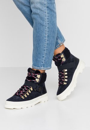 Person, der blaue Jeans und marineblaue Wanderschuhe mit roten und weißen Schnürsenkeln, goldenen Ösen und weißen, klobigen Sohlen trägt, auf einem einfarbigen Hintergrund.