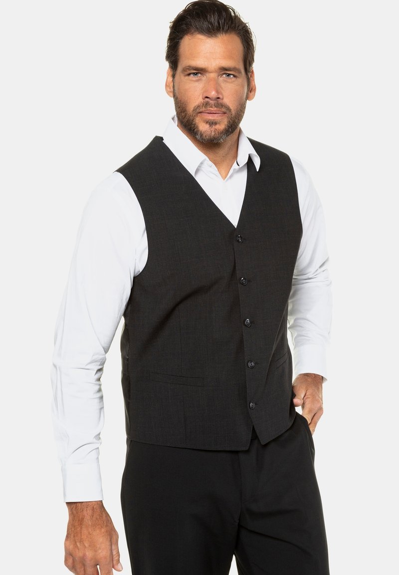 Chaleco negro con estampado y cinco botones, llevado sobre una camisa blanca, con escote en pico y bolsillos laterales, combinado con pantalones negros.