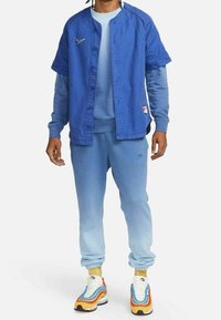 Modrá oversized košile s knoflíky a kulatým límcem přes světle modrou mikinu a odpovídající tepláky. Barevné tenisky se vrstveným designem.