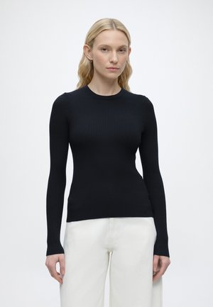 Hosszú, szőke hajú nő fekete testhez simuló bordázott pulóvert és fehér nadrágot visel egy egyszínű világos háttér előtt, előre nézve áll.