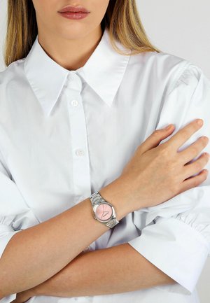Silberne Uhr mit rosa Zifferblatt, Metallarmband und minimalistischem Design. Am Handgelenk getragen, kombiniert mit einem weißen Hemd mit Kragen.