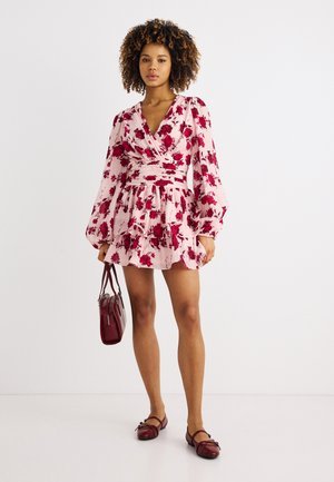 Žena stojaca v ružových kvetinových šatách zavinovacieho strihu s dlhými rukávmi, držiaca červenú kabelku a obutá v červených balerínkach Mary Jane.