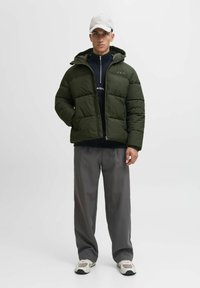 Grüne Pufferjacke mit Kapuze, die ein gestepptes Design aufweist. Getragen über einem schwarzen Reißverschluss-Oberteil und kombiniert mit grauen Hosen und Sneakers.