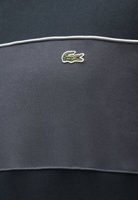 Tissu gris foncé et noir avec un liseré blanc et un petit logo de crocodile vert brodé centré sur la partie supérieure.