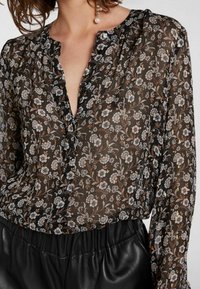 Zwarte doorschijnende blouse met bloemendessin in wit, beige en groen. Heeft een diepe V-hals en een gerimpelde sluiting. Gecombineerd met zwarte gestructureerde broek.