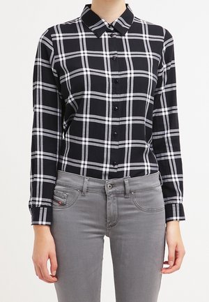 Schwarz-weiß kariertes Hemd mit Knöpfen, getragen mit hellgrauen Jeans bei einer Person, die vor einem einfarbigen Hintergrund steht.
