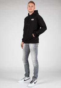 Felpa nera con tasca frontale, cappuccio con coulisse e dettaglio del logo colorato. Abbinata a jeans grigio chiaro e scarpe da ginnastica bianche con accenti neri.