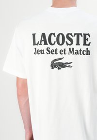Balta īsām piedurknēm T-kreklā ir melns teksts "LACOSTE Jeu Set et Match" un melns krokodila grafiks aizmugurē. Izgatavots no kokvilnas auduma.
