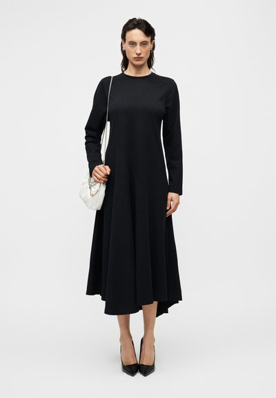 Μακρυμάνικο μαύρο φόρεμα με φουσκωτή σχεδίαση, που διαθέτει στρογγυλή λαιμόκοψη και μήκος μέχρι το γόνατο, συνδυασμένο με λευκή καπιτονέ τσάντα και μαύρες γόβες.