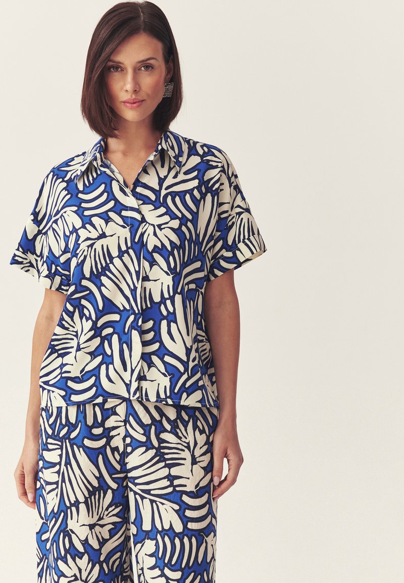 Femme aux cheveux bruns courts portant une chemise à manches courtes et un pantalon assortis à imprimé tropical bleu et blanc, debout devant un fond uni.