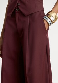 Vyno raudonos spalvos pritaikytos kelnės su tekstūruotu paviršiumi, turinčios šoninę kišenę ir formalų sagos detalę juosmens srityje, derinamos su sutvirtintomis apyrankėmis.