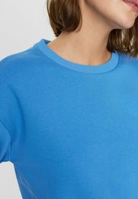 Mėlynas džemperis su smulkiais krūtinės raštai, pagamintas iš minkštos medžiagos, turintis lygią tekstūrą ir laisvą siluetą.