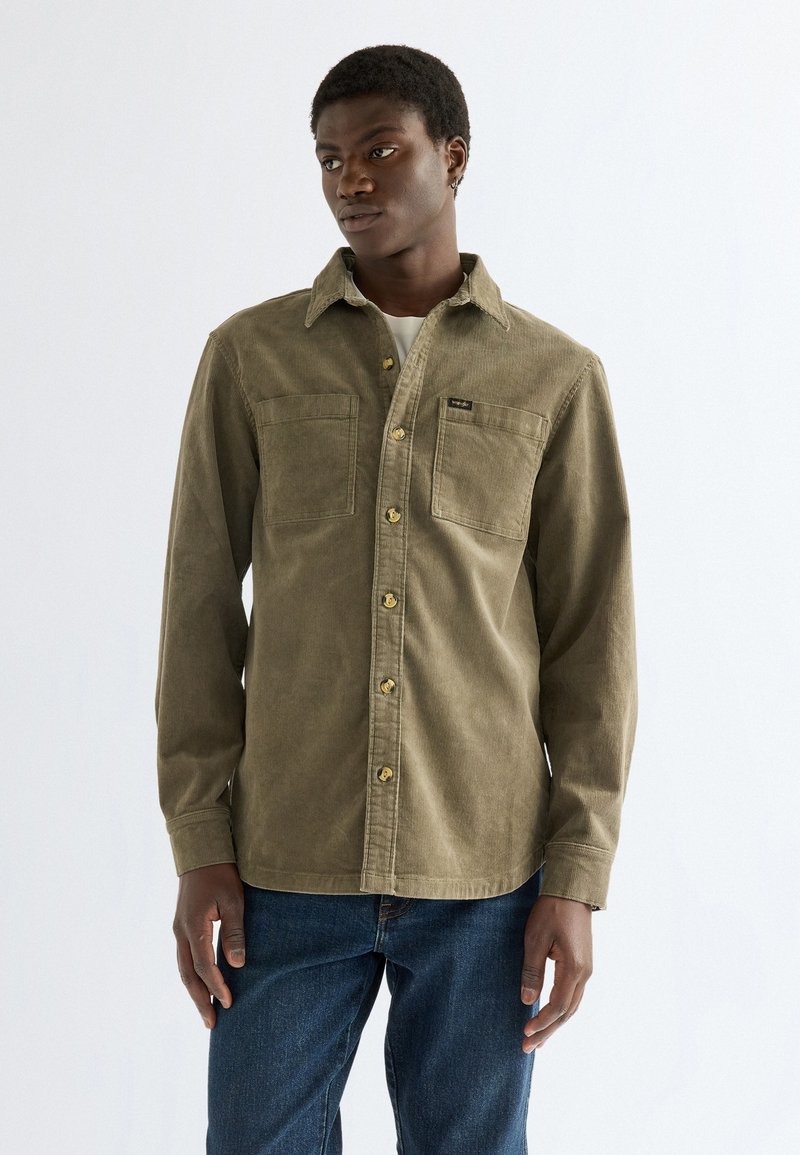 Jeune homme portant une chemise en velours côtelé vert olive à boutons avec deux poches poitrine et un jean bleu, debout devant un fond blanc uni.