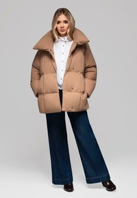 Kort, beige pufferjakke med en bred krave, store quiltede sektioner og knapper. Den er parret med en hvid skjorte og mørkeblå vide jeans.