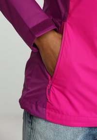 Main d'une personne dans la poche latérale zippée d'une veste violette et rose, avec un détail de poignet en velcro et un jean en denim bleu clair.