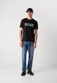 Schwarzes Baumwoll-T-Shirt mit "BOSS" in Weiß bedruckt, kombiniert mit hellblauen Jeans und schwarzen Sneakers. Eine schwarze Baseballmütze rundet das Outfit ab.