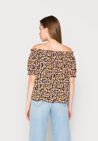 Blusa off-the-shoulder em tecido preto, com pequenas flores amarelas e cor-de-rosa, mangas bufantes e decote elástico, combinada com jeans claros.
