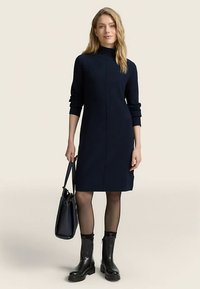 Tmavomodré dlhé šaty s dlhým rukávom, vysokým golierom a rovým strihom. Nosené s čiernymi čižmami dĺžky do polovice lýtok a drží čiernu kabelku.