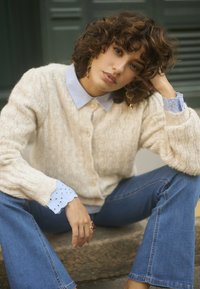Cardigan en tricot torsadé crème avec des boutons en perles, superposé à une chemise à col bleu clair. Accents en eyelet bleu clair sur les manches, associé à un jean bleu.