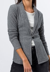 Veste légère grise à rayures avec un col châle, une fermeture à un bouton et des manches longues, portée sur une chemise blanche et un jean noir.