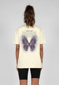 Camiseta de color amarillo claro con un gran gráfico de una mariposa morada en la parte posterior, acompañado de texto negro en la parte superior. Mangas cortas y corte relajado.