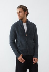 Cardigan en tricot gris zippé avec un tissu texturé, deux poches avant et des poignets côtelés. Porté par-dessus un t-shirt blanc, avec une coupe décontractée.