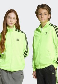 Neongröna Adidas-trackjackor med svarta ränder, dragkedja framtill och logotyp på bröstet; båda bärs av en flicka och en pojke, kombinerade med lediga byxor.
