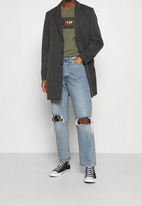 Abrigo gris ajustado sobre camiseta oliva con diseño gráfico, combinado con jeans azul claro desgastados y zapatillas negras de caña alta.