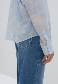 Camicia trasparente azzurro chiaro con un sottile motivo bianco, maniche lunghe, polsini con un solo bottone, abbinata a pantaloni in denim blu con una tasca posteriore.