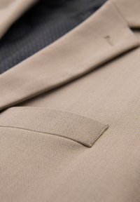 Veste de costume beige à motif chevrons avec un gros plan du revers cranté et de la poche passepoilée.