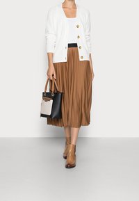 Cardigan bianco a maglia con bottoni dorati, gonna plissettata marrone, stivali alla caviglia color tan e borsa tote nera con una tasca frontale beige.