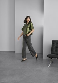 Groen pailletten top met brede mouwen, gecombineerd met grijze wijde jeans en zwarte hakken. Achtergrond met grijze muren en meubels.