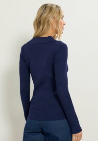 Marinblå ribbad tröja med krage, långa ärmar och en figurnära passform. Mjuk textur visad från en bakvinkel, parat med jeans.