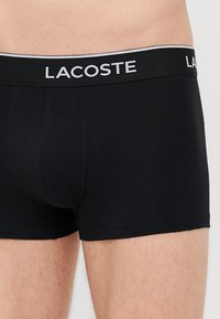 Crne bokserice od pamuka s uskim krojem, s belim pojasom s logotipom na kojem piše "LACOSTE" velikim slovima.