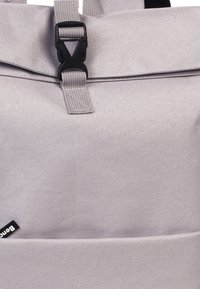 Grijze stoffen rugzak met een klep-sluiting, zwart metalen clip en een rechthoekige silhouette. Inclusief een klein merkembleem.