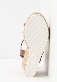 Sandale compensée avec semelle blanche rainurée, semelle intermédiaire tissée de style espadrille et brides en cuir marron appuyées contre un mur blanc.