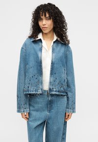 Ανοιχτό μπλε τζιν μπουφάν με κομψή cropped σιλουέτα, διακοσμημένο με μεταλλικά τρουκς, διαθέτει κλασικό γιακά και δύο πλαϊνές τσέπες.